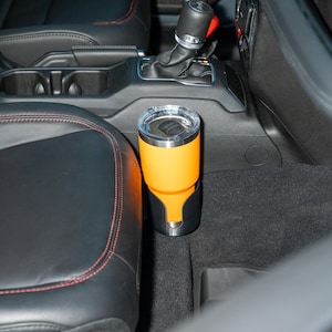 May include: An orange insulated tumbler with a silver rim and a clear lid sits in a black car cup holder. The cup holder is designed to fit snugly in a vehicle's console. The interior of the car features black leather seats with red stitching.