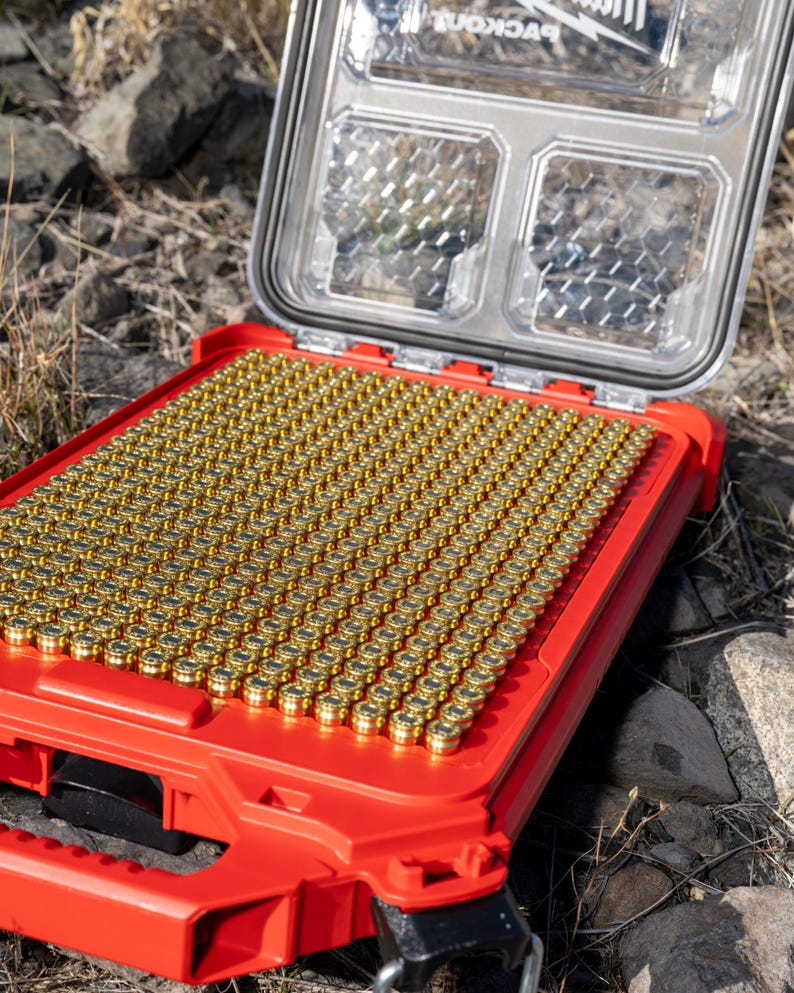May include: Open red storage case filled with numerous gold-colored bullet casings. The clear lid is open, revealing the contents. The case is on a rocky surface.