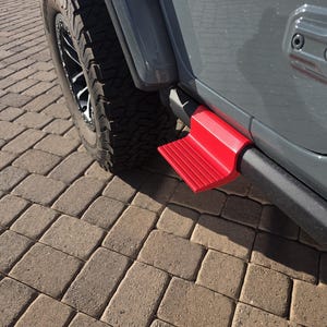May include: A bright red, textured step accessory mounted to the side of a gray vehicle. The step features a ridged surface for traction and is positioned below the door frame, adjacent to a black tire with a rugged tread pattern on a paved surface.