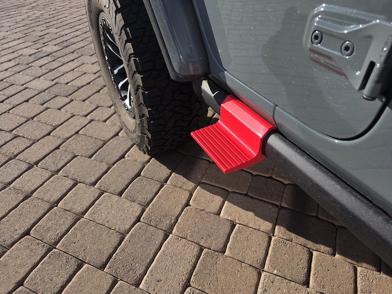 May include: A bright red, textured step accessory mounted to the side of a gray vehicle. The step features a ridged surface for traction and is positioned below the door frame, adjacent to a black tire with a rugged tread pattern on a paved surface.