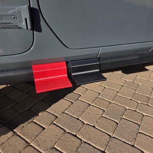 May include: A pair of exterior vehicle door steps attached to the lower frame of a gray car. One step is bright red and the other is matte black. Both steps feature a horizontal ribbed texture on the surface to provide grip for stepping.