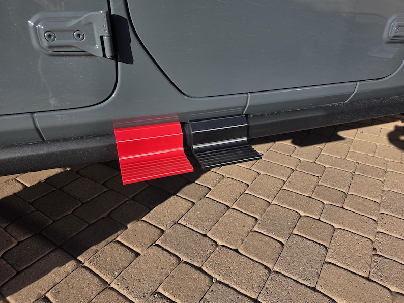 May include: A pair of exterior vehicle door steps attached to the lower frame of a gray car. One step is bright red and the other is matte black. Both steps feature a horizontal ribbed texture on the surface to provide grip for stepping.