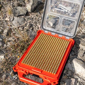 Puede incluir: Una caja de almacenamiento rectangular naranja abierta, llena de numerosos objetos cil&iacute;ndricos peque&ntilde;os de color dorado. La caja tiene una tapa transparente con m&uacute;ltiples compartimentos. La marca "Milwaukee" es visible.