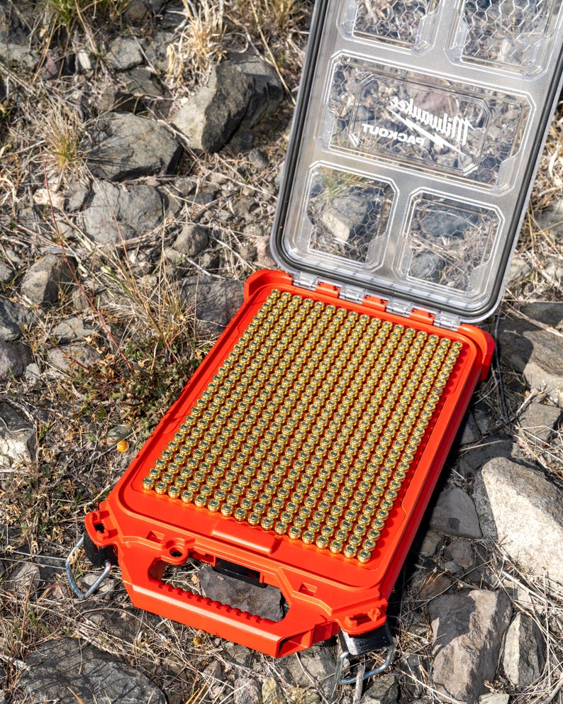 Puede incluir: Una caja de almacenamiento rectangular naranja abierta, llena de numerosos objetos cil&iacute;ndricos peque&ntilde;os de color dorado. La caja tiene una tapa transparente con m&uacute;ltiples compartimentos. La marca "Milwaukee" es visible.