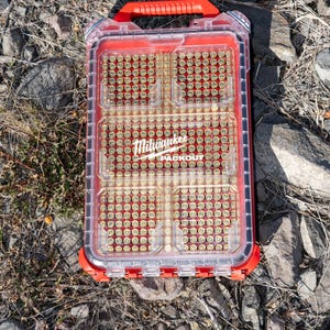 Puede incluir: Un contenedor de almacenamiento Milwaukee Packout transparente y rojo, lleno de peque&ntilde;os objetos de color lat&oacute;n. El contenedor tiene un asa roja y descansa sobre un lecho de rocas y grava. El logotipo de Milwaukee es visible.