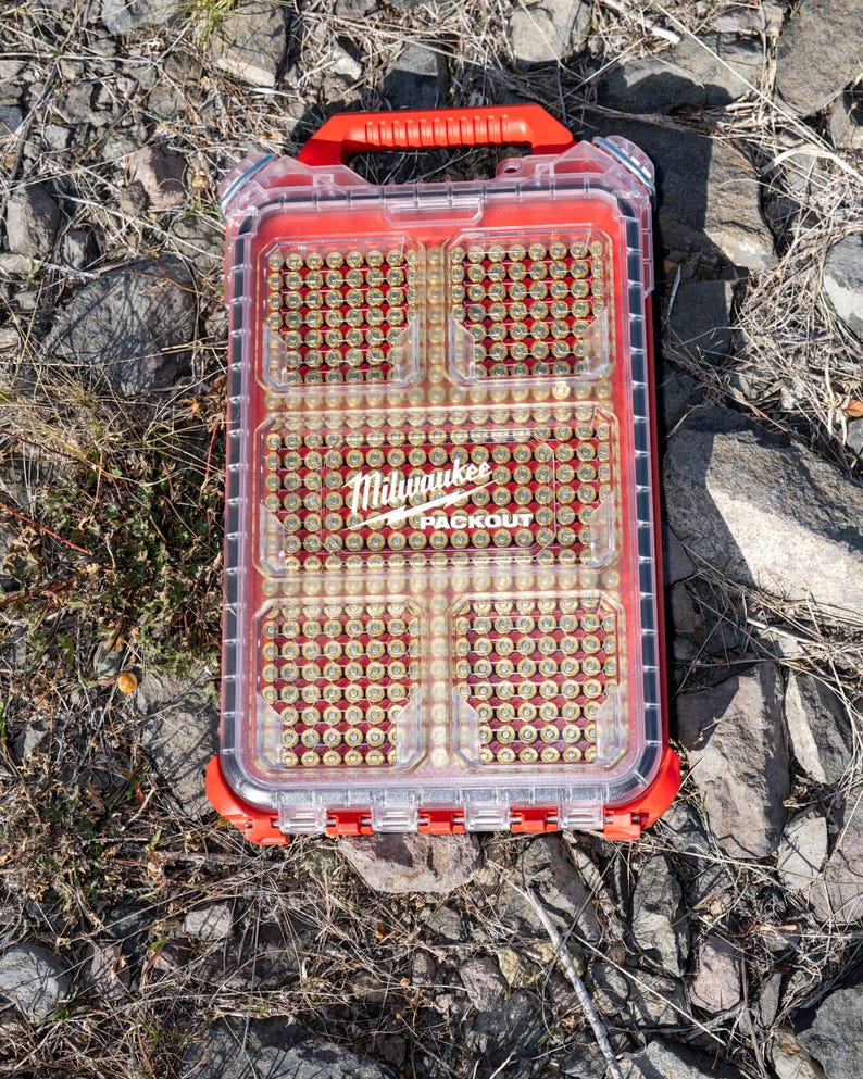 Puede incluir: Un contenedor de almacenamiento Milwaukee Packout transparente y rojo, lleno de peque&ntilde;os objetos de color lat&oacute;n. El contenedor tiene un asa roja y descansa sobre un lecho de rocas y grava. El logotipo de Milwaukee es visible.