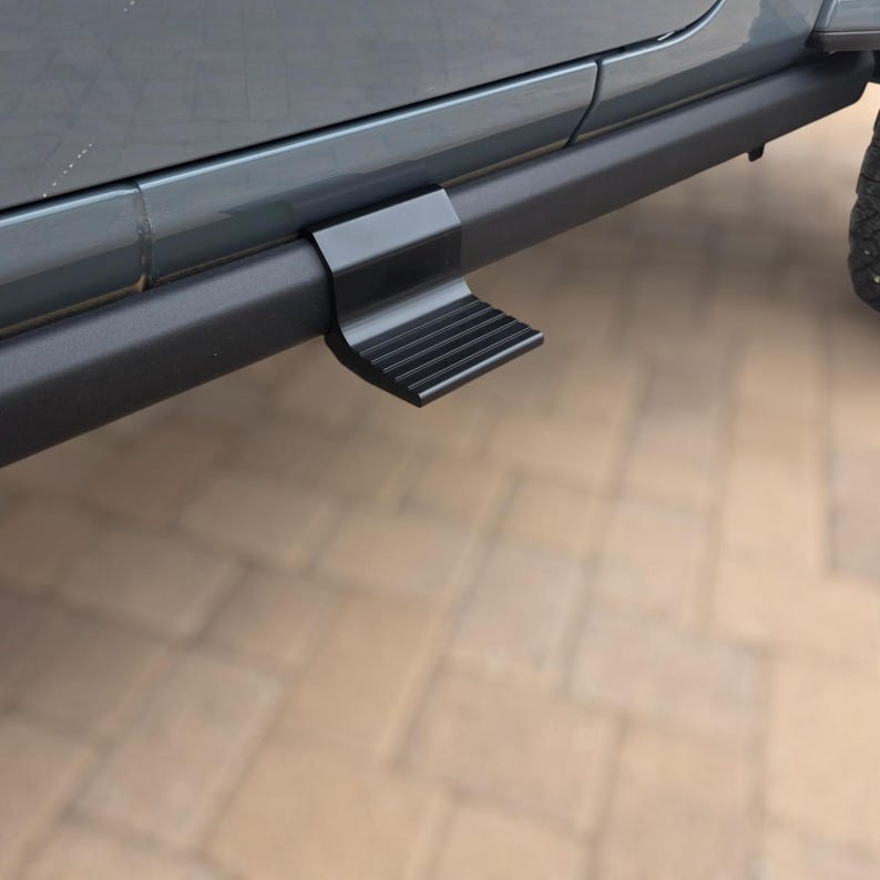 May include: A matte black metal vehicle door step accessory attached to the side rocker panel of a gray vehicle. The step features a rectangular design with horizontal ridges on the surface to provide traction for stepping into or out of the vehicle.