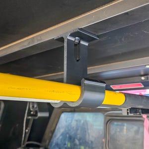 May include: A black metal mounting bracket secured to a horizontal surface, featuring a U-shaped cradle that holds a long, cylindrical yellow pole. The bracket is attached with a visible screw and provides a stable support for the yellow tool handle.