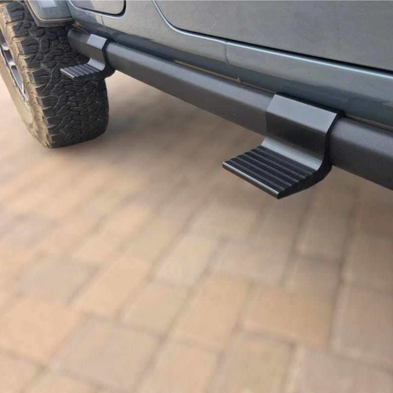 May include: A pair of matte black metal side steps mounted to the lower rocker panel of a gray vehicle. The steps feature a textured, ribbed surface for traction and are positioned horizontally along the side, extending outward from the vehicle body.