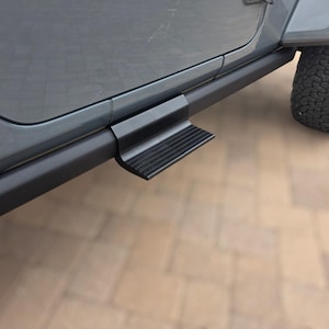 May include: A black metal side step mounted to the lower rocker panel of a gray vehicle. The step features a rectangular, textured surface with horizontal ridges designed for traction, extending outward from the vehicle's side frame.