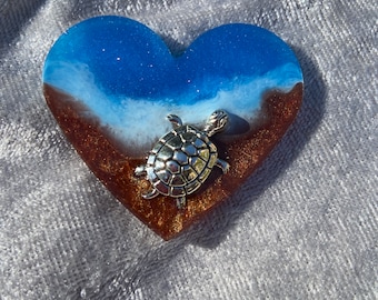 May include: A heart-shaped resin ornament with a blue and white ocean scene and a silver turtle on top. The ornament has a brown sand-like base.