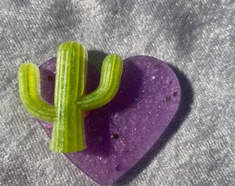 May include: A purple heart-shaped dish with glitter embedded in the resin. A green cactus is sitting on top of the dish.