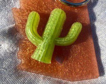 May include: A pink glitter resin keychain shaped like the state of Arizona with a green cactus in the center.