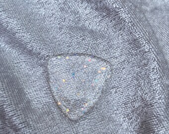 May include: A clear, heart-shaped resin charm with iridescent glitter embedded inside. The charm is lying on a gray, textured fabric.