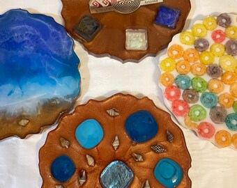 May include: Four resin coasters with different designs. One coaster features a blue and white ocean wave design. Another coaster has a brown background with blue and iridescent circles. A third coaster has a brown background with a turquoise and white geometric design. The fourth coaster has a brown background with a turquoise and white geometric design and the text "Arizona" and "Navajo" in gold.