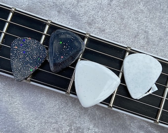 May include: Four guitar picks on a guitar fretboard. The picks are black with glitter, white with glitter, and white with a marble pattern.
