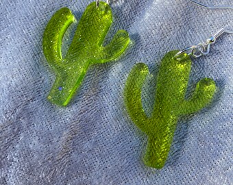 May include: A pair of green glitter cactus earrings with silver hooks. The earrings are made of resin and have a sparkly finish.