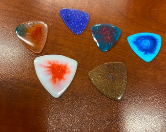 May include: Six different guitar picks with various colors and patterns. The picks are made of clear plastic with glitter embedded in the plastic. The colors include blue, red, orange, white, and brown.