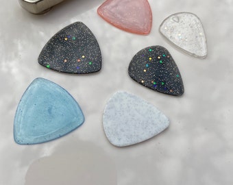 May include: Six guitar picks with different glitter patterns. The picks are in the shape of a triangle and are made of a clear material. The colors of the glitter are pink, black, white, blue, and silver.