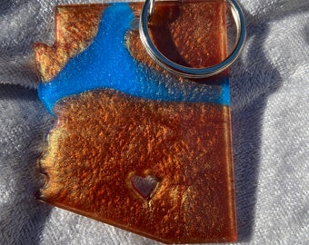 May include: A copper-colored Arizona state keychain with a blue river design and a heart cutout. The keychain has a silver key ring attached. The resin has a glittery, shimmering effect.