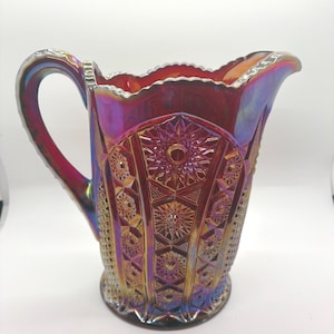 Puede incluir: Una jarra de vidrio iridiscente rojo rubí con un patrón decorativo de estrellas y formas geométricas. La jarra tiene un asa y un borde festoneado. El vidrio refleja colores púrpura, azul y dorado.