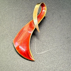 Peut inclure: Une broche en émail rouge et or avec un design abstrait incurvé. La broche est en métal et a une finition brillante.