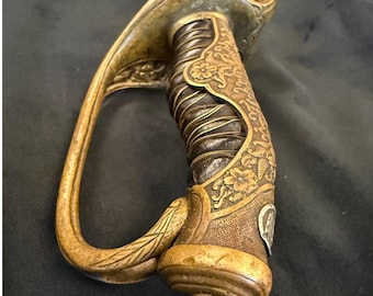 May include: Close-up of an antique sword hilt. The hilt is gold-toned with intricate floral and scrollwork designs. The grip is wrapped in dark brown leather and wire. The guard curves gracefully, adding to the ornate aesthetic.