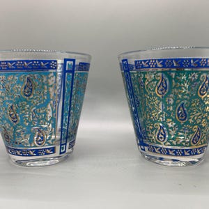May include: Two clear glass tumblers with a turquoise and gold paisley design. The glasses have a blue border and a decorative pattern around the rim. The tumblers are set against a white background.
