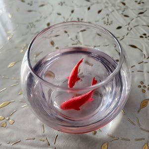 Op de afbeelding: Twee rode plastic goudvisjes in een glazen kom op een witte tafelkleed met een gouden patroon.