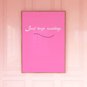 Puede incluir: Un estampado artístico rosa enmarcado con las palabras "Just keep reading" en escritura blanca. Una línea negra curva está debajo del texto. El marco es de color marrón claro. El estampado está colgado en una pared rosa.