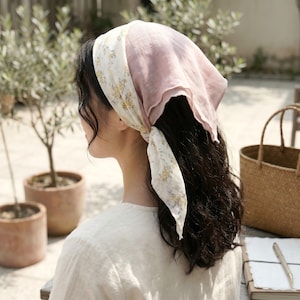 Puede incluir: Un pañuelo para la cabeza con un estampado floral y una sección rosa liso. El pañuelo está atado en la nuca, con la sección estampada con pequeñas flores amarillas sobre un fondo crema. La sección lisa es rosa suave.