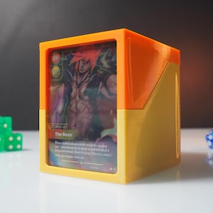 May include: A two-toned orange and yellow plastic deck box featuring a clear front window displaying a trading card with an illustration of a character and the text The Boss. Three green dice and one blue die are positioned near the box.