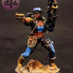 May include: Miniature figurine of a female character in a futuristic outfit, holding a pistol and a large weapon. The figure is painted with detailed shading and features a blue top, brown jacket, and blue pants. The base is textured with sand and small rocks.