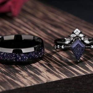 May include: A black wedding band with a dark blue, sparkly finish and a black engagement ring with a large, dark blue, sparkly centre stone and smaller, clear stones around it.