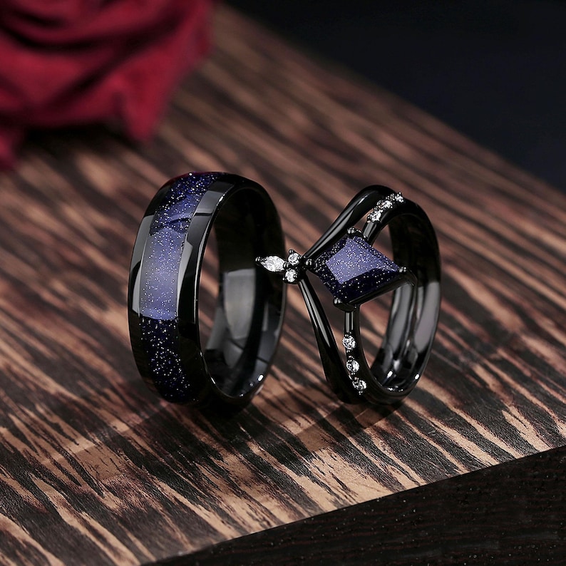 May include: A pair of black wedding bands with a blue starry sky design. The woman's band features a square-shaped blue stone with smaller clear stones on the band.