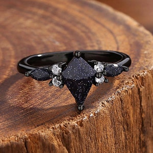 May include: Black ring with a large, square-shaped, dark blue gemstone surrounded by smaller, white gemstones. The ring band is black.