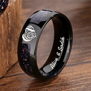 Puede incluir: Anillo de carburo de tungsteno negro con incrustaciones de purpurina negra y morada. El anillo está grabado con el texto "Alice & Sadik" y un corazón estilizado con la letra "A" en su interior.