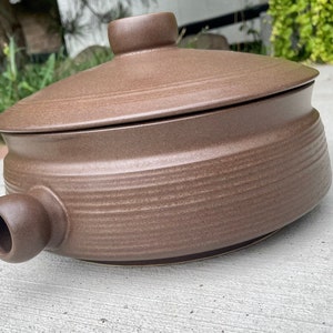 May include: A brown ceramic pot with a lid and a spout. The pot has a textured surface and is designed for cooking.