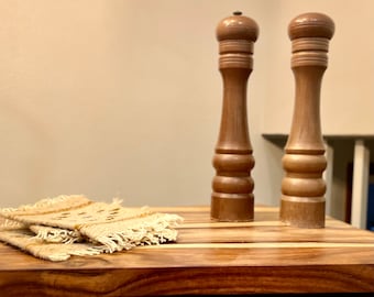 Può includere: Due macine per sale e pepe in legno su un tagliere di legno con un sottopentola in tessuto.