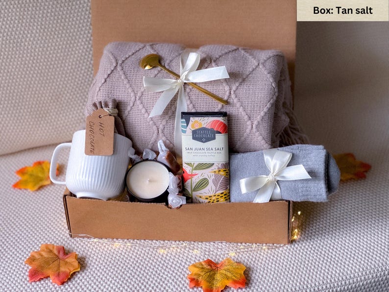 May include: A gift box filled with cozy items for a relaxing evening. It includes a soft blanket, a mug that says "Hot Chocolate", a candle, a bar of chocolate, and a pair of socks. The box is tan.