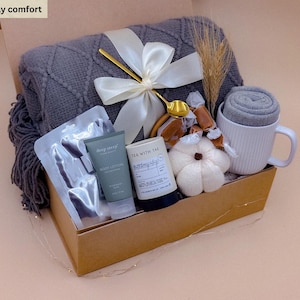 Könnte beinhalten: Eine Geschenkbox mit der Aufschrift Sunday comfort enthält eine dunkelgraue Strickdecke mit weißem Satinband, einen goldenen Löffel, eine weiße gerippte Tasse mit grauen Socken, eine Körperlotion, eine Kerze mit der Aufschrift Tea with Tae und einen weißen Kürbis.