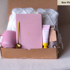 May include: A pink and gold gift box filled with items for a self-care night. The box contains a pink mug, a pink notebook, a gold spoon, a tube of hand cream, and a small bag of hot chocolate.