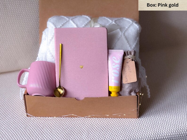 May include: A pink and gold gift box filled with items for a self-care night. The box contains a pink mug, a pink notebook, a gold spoon, a tube of hand cream, and a small bag of hot chocolate.