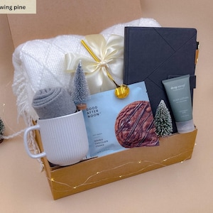 Könnte beinhalten: Eine Geschenkbox mit der Aufschrift Glowing Pine enthält eine weiße Strickdecke, eine graue Tasse, ein graues Notizbuch, eine Tube Körperlotion, eine Packung Good After Noon Doppelschokoladenkekse, einen goldenen Löffel und kleine Deko-Tannen.
