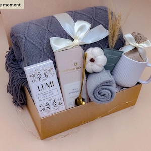 Könnte beinhalten: Eine Geschenkbox mit der Aufschrift Box: Gentle moment, die eine graue Strickdecke, eine weiße Tasse, eine Lumi weiße heiße Schokolade, eine Moonstruck Milchschokolade, einen goldenen Löffel, einen kleinen weißen Kürbis und einen Beutel enthält.