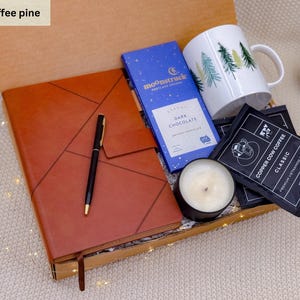 K&ouml;nnte beinhalten: Eine Geschenkbox gef&uuml;llt mit einem braunen Lederjournal, einem schwarzen Stift, einer blauen Schokoladentafel, einer wei&szlig;en Tasse mit gr&uuml;nen B&auml;umen, einer schwarzen Kerze und Kaffeep&auml;ckchen. Die Box ist offen.