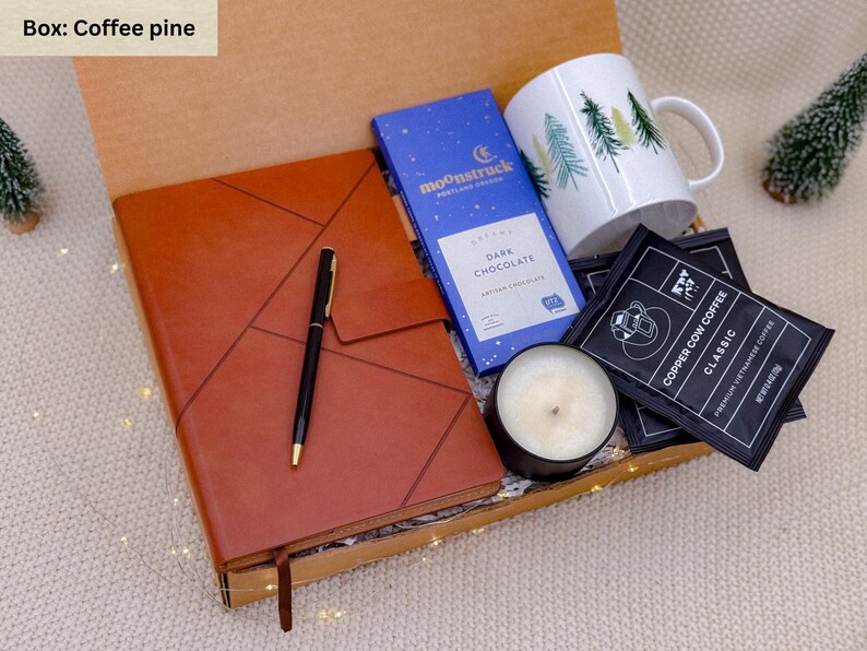 K&ouml;nnte beinhalten: Eine Geschenkbox gef&uuml;llt mit einem braunen Lederjournal, einem schwarzen Stift, einer blauen Schokoladentafel, einer wei&szlig;en Tasse mit gr&uuml;nen B&auml;umen, einer schwarzen Kerze und Kaffeep&auml;ckchen. Die Box ist offen.
