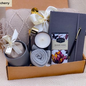 K&ouml;nnte beinhalten: Geschenkbox in Beige mit einem grauen Notizbuch, Stift, Kerze, Schokoriegel und einer kleinen gerollten Decke. Ein beiges Kissen und ein kleiner Beutel mit wei&szlig;em Band sind ebenfalls enthalten.