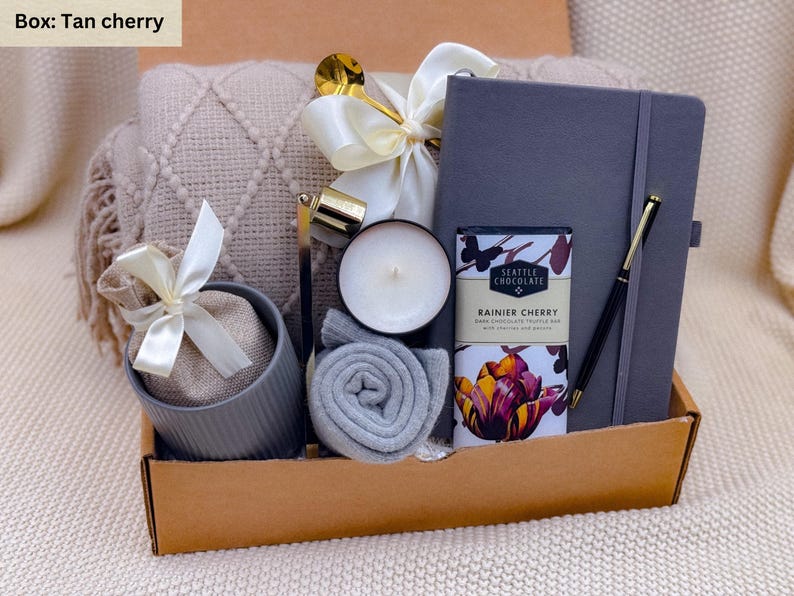 K&ouml;nnte beinhalten: Geschenkbox in Beige mit einem grauen Notizbuch, Stift, Kerze, Schokoriegel und einer kleinen gerollten Decke. Ein beiges Kissen und ein kleiner Beutel mit wei&szlig;em Band sind ebenfalls enthalten.