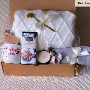 May include: A gift box filled with a white blanket, mug, Seattle Chocolate bar, candle, and gray socks tied with a white ribbon. The mug says "I love you" with red hearts. A gold spoon rests on the blanket.